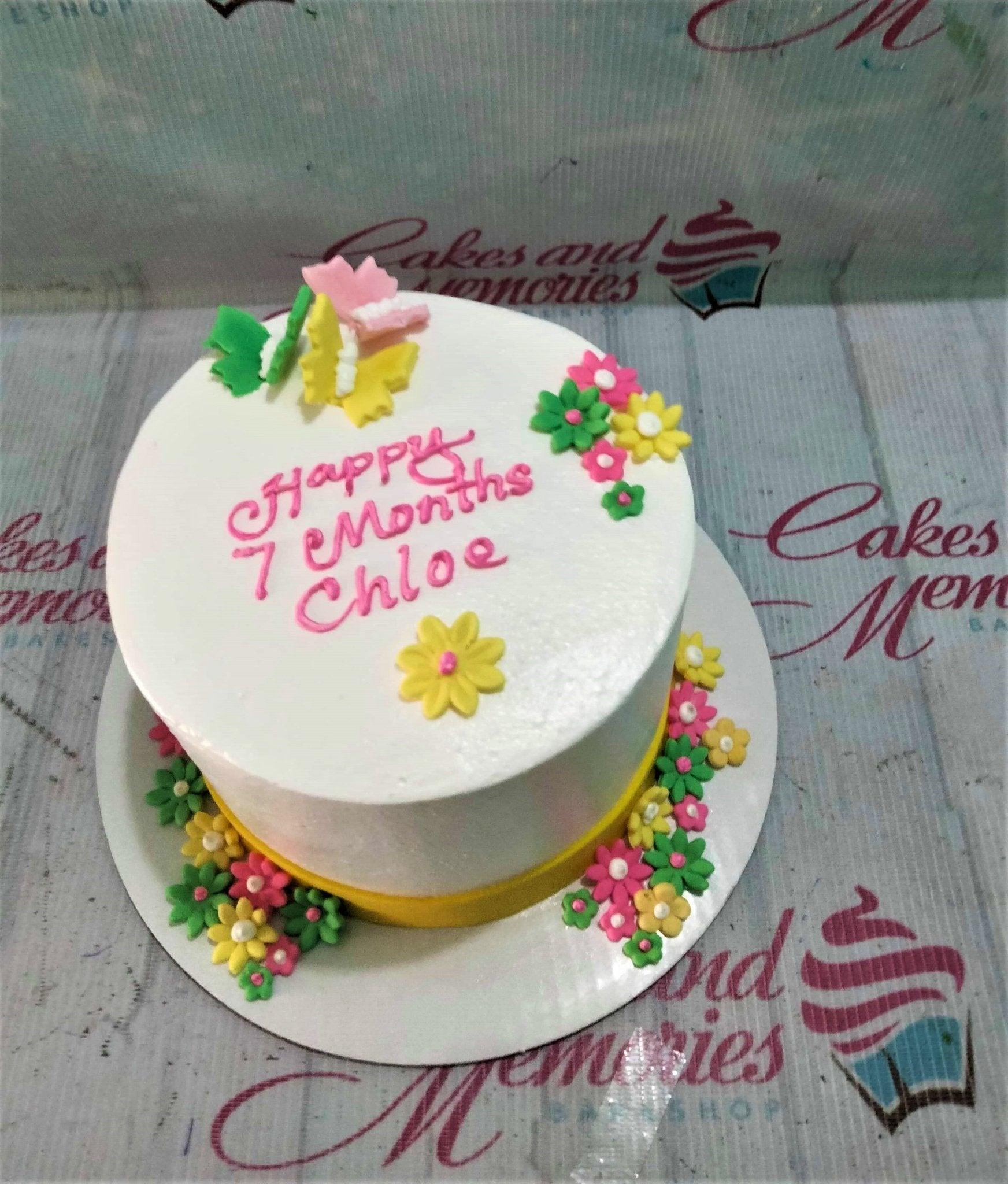 White round cake with pink icing message, yellow and pink fondant flowers, and colorful fondant butterflies.