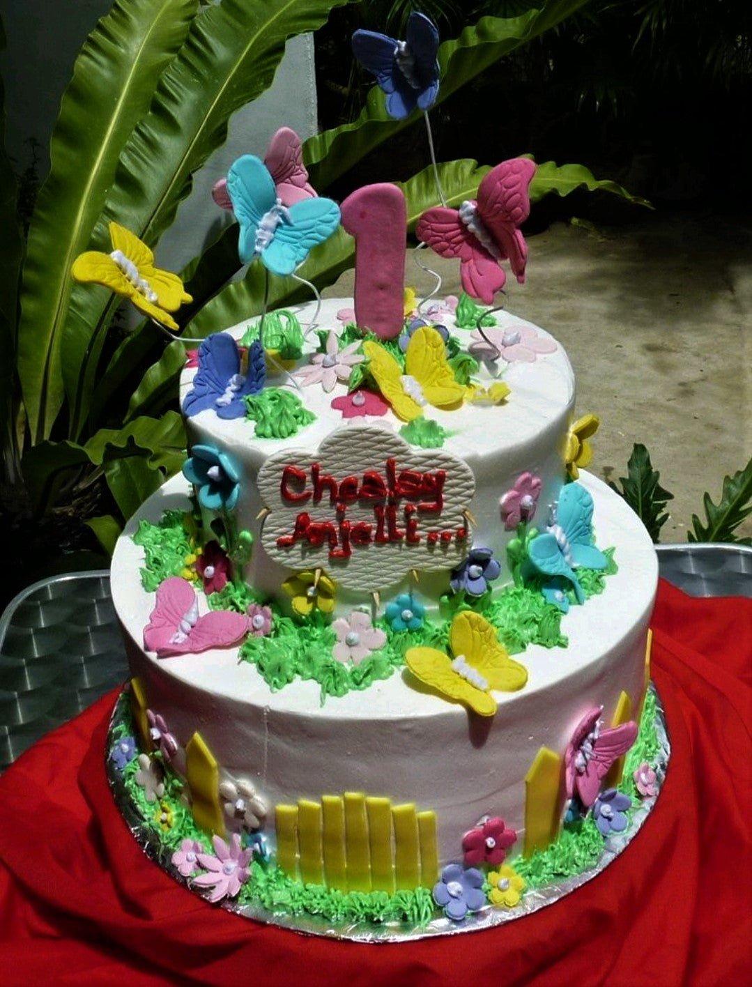 Two-tier garden-themed birthday cake with colorful fondant butterflies, flowers, a yellow fence, and a pink number one topper.