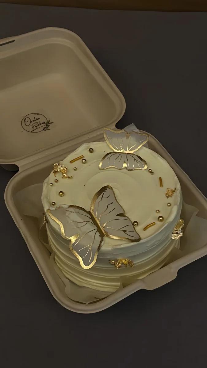 White bento cake with gold-edged butterfly toppers, gold leaf accents, and metallic gold sprinkles in a compostable box.