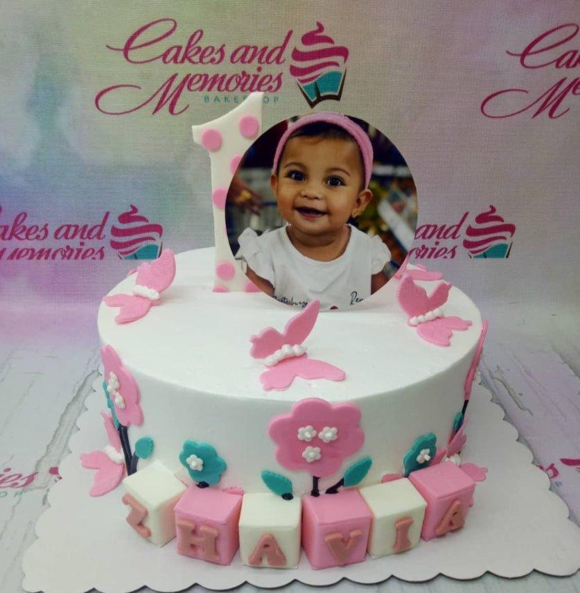 White 1-tier birthday cake with pink butterflies, flowers, and a photo printout topper for a first birthday celebration.