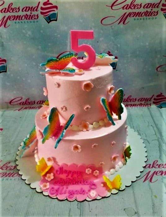 Pink two-tier butterfly birthday cake with rainbow butterfly printouts and small pink fondant flowers.
