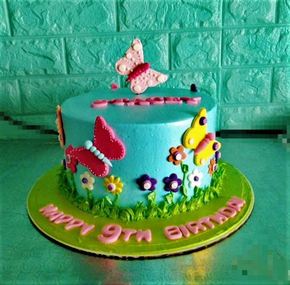 Light blue butterfly birthday cake with pink, yellow, and purple fondant butterflies and flowers with green icing grass border.