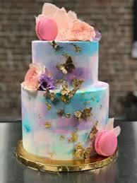 A soft blue 2 tier cake featuring gold butterflies, pink macarons, peach edible flowers, a purple flower, and pink sugar shards.