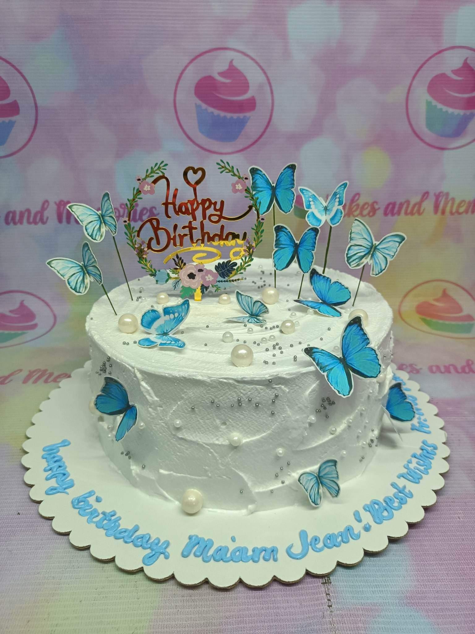 White soft-icing butterfly cake with blue printout butterflies, pearls, and a gold Happy Birthday topper.