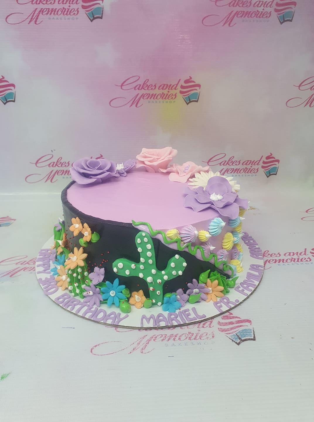 A round purple and black cake decorated with fondant flowers, a green cactus, and a birthday message on the base board.