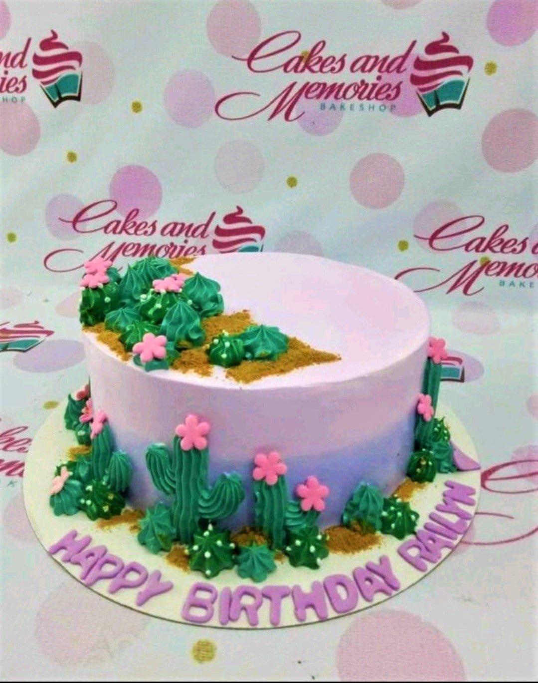 A purple and light blue gradient cake decorated with green piped icing cacti and pink fondant flowers on a white board.