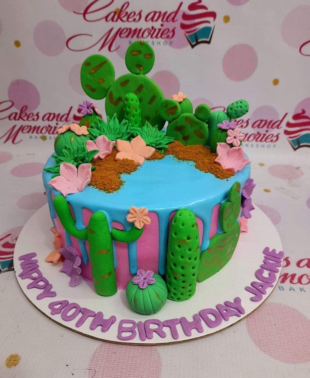 Cactus themed birthday cake with pink and blue drip icing, fondant cacti, and purple gumpaste letters on the base board.