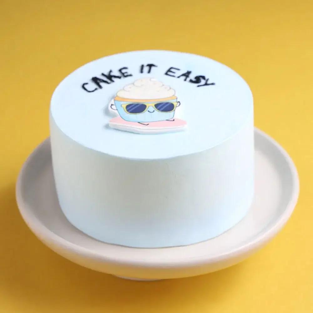 Light blue bento cake with a cute cupcake character wearing sunglasses and the message 'CAKE IT EASY' in black icing.