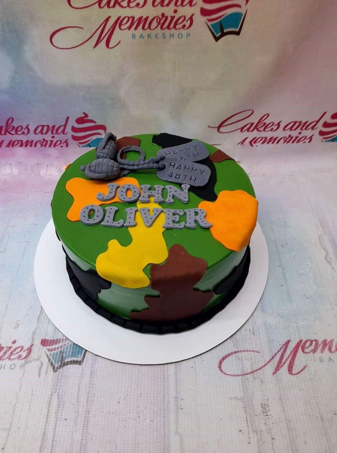 Camouflage military themed cake with green, brown, yellow, and black fondant patches, featuring a fondant grenade and dog tags.