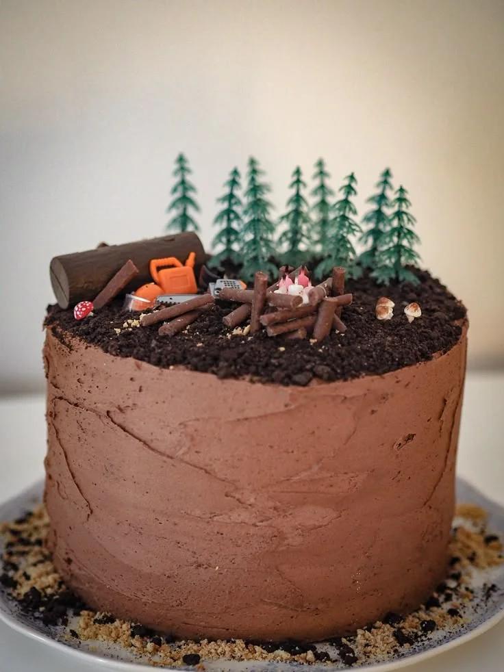 Chocolate camping themed cake with a forest of pine trees, a campfire made of chocolate logs, and a chainsaw topper.