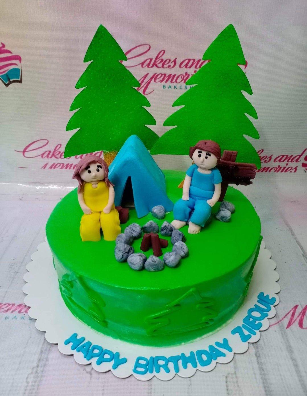 Green camping themed birthday cake with fondant figures of a couple, a blue tent, campfire, and pine trees.