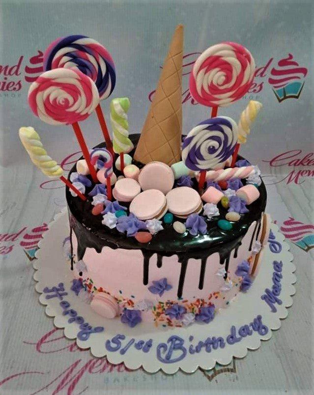 Pink birthday cake with chocolate drip, colorful lollipops, marshmallows, macarons, and purple icing rosettes.