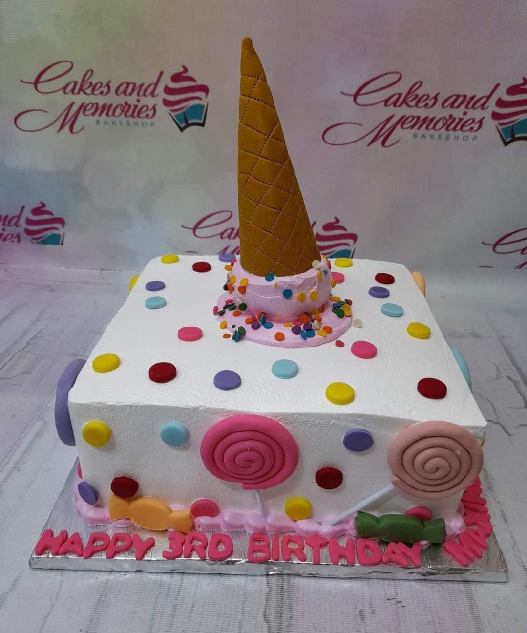 Square white soft-icing cake with colorful fondant dots, lollipops, candies, and a large ice cream cone topper.