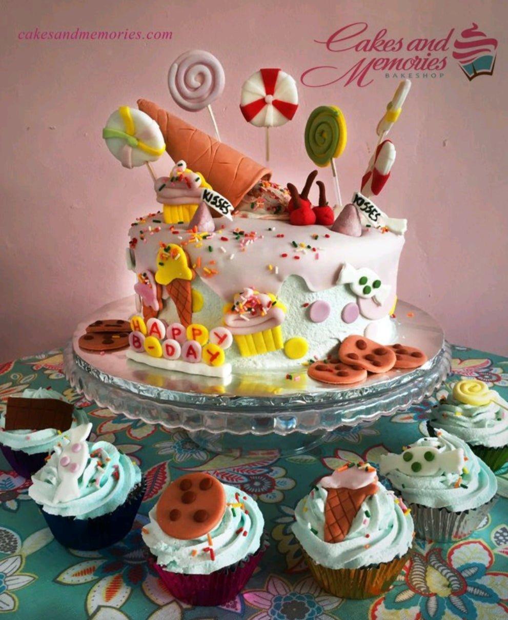 A whimsical candy-themed birthday cake with a pink and light blue icing base, topped with an upside-down ice cream cone and various sweets.