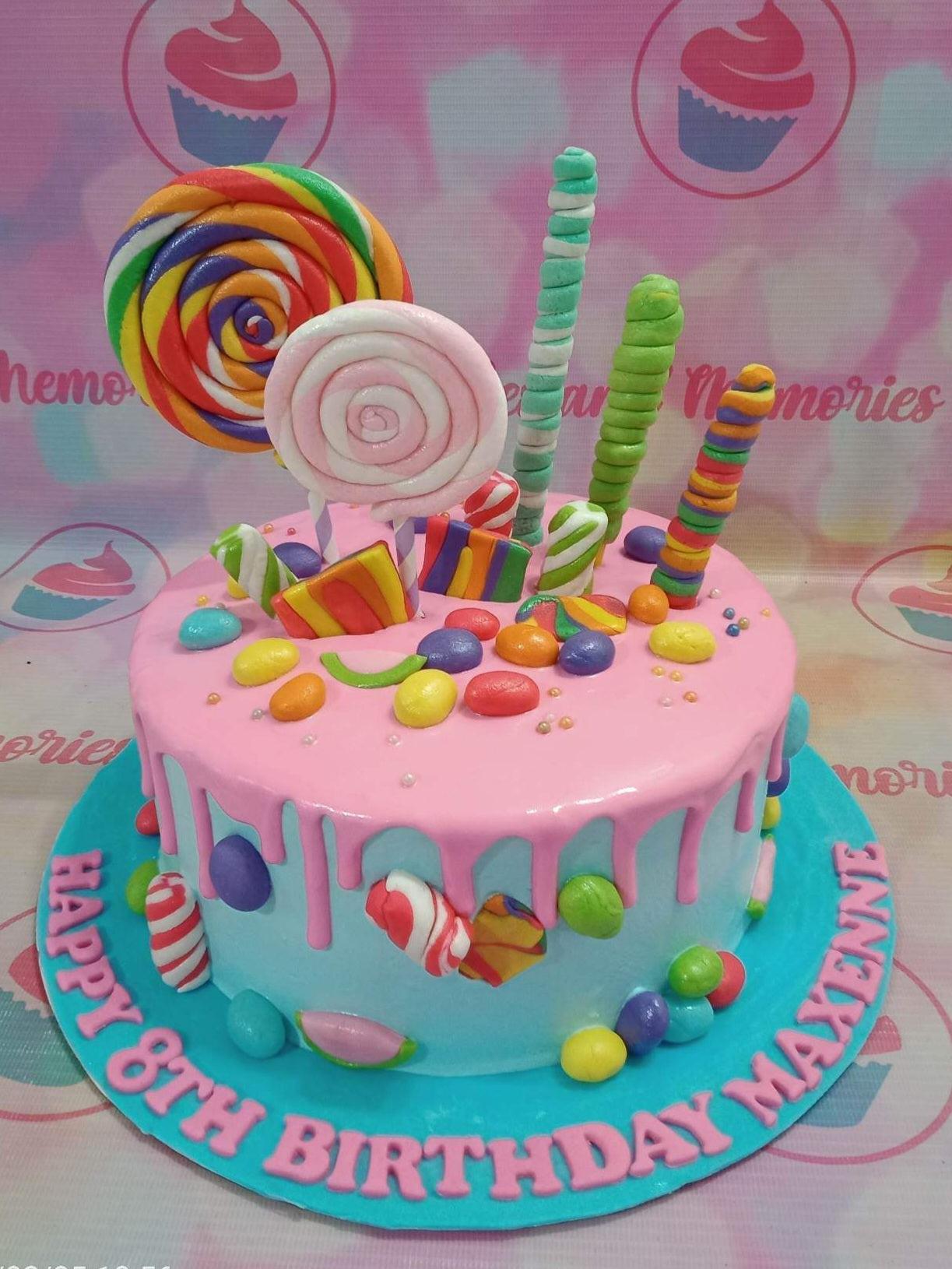 Colorful candy-themed birthday cake with pink drip icing, lollipops, and assorted fondant sweets on a light blue base.