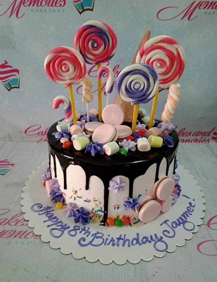 Pink birthday cake with chocolate drip, colorful lollipops, marshmallows, and macarons on a white base board.