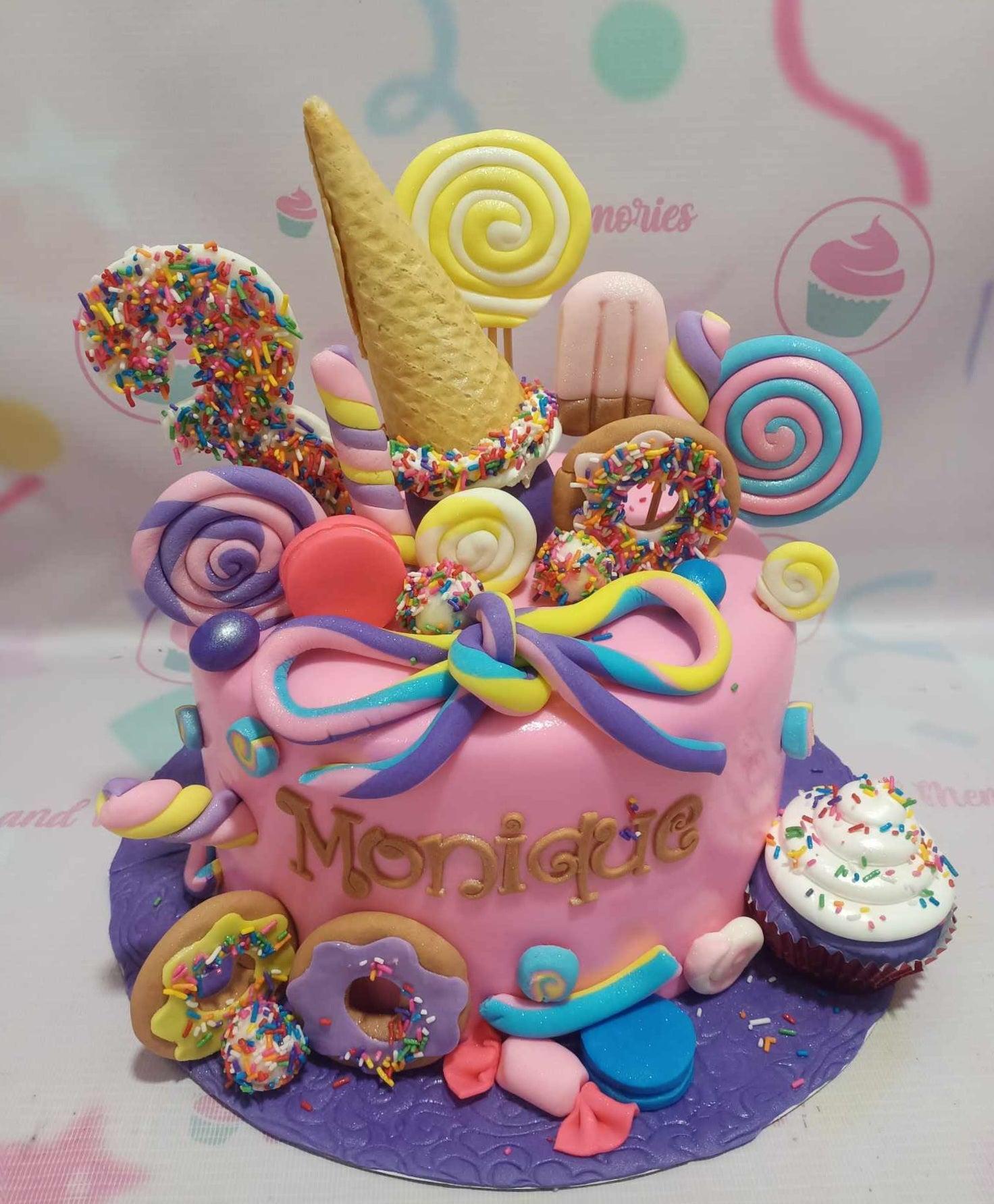 Pink candy-themed fondant cake with lollipops, donuts, ice cream cone, and a cupcake on a purple base.