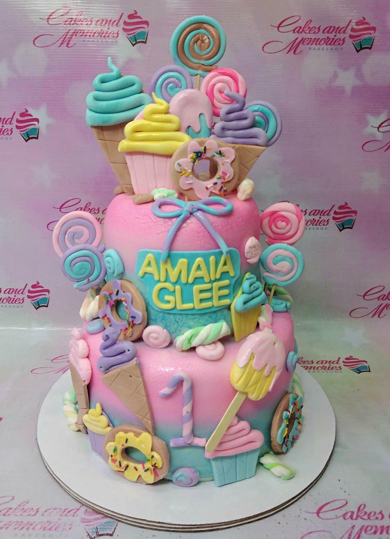 Colorful 2-tier candy themed birthday cake with fondant ice cream cones, donuts, lollipops, and cupcakes in pastel pink, blue, and yellow.