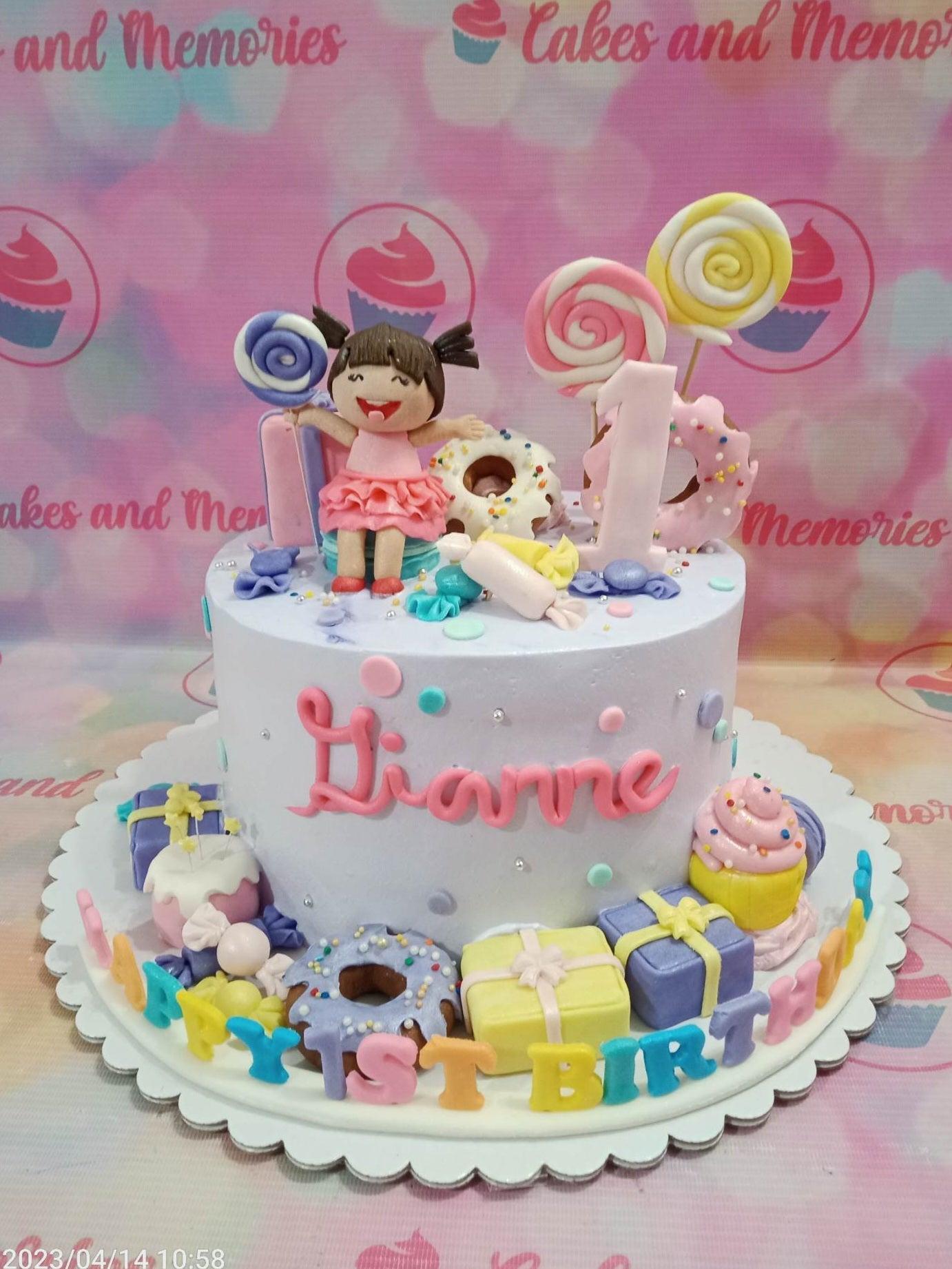Light purple 1-tier birthday cake with a fondant girl figure, donuts, lollipops, and colorful gift boxes on a white base board.