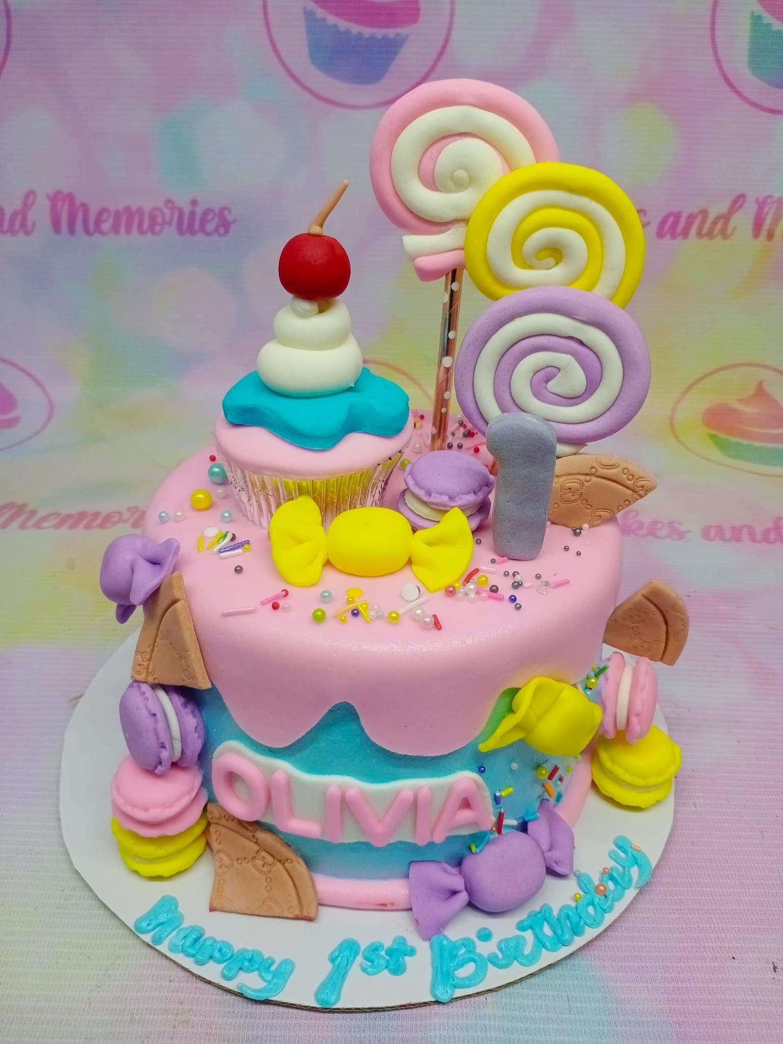Colorful candy-themed birthday cake with pink and blue icing, topped with a fondant cupcake, lollipops, and macarons.