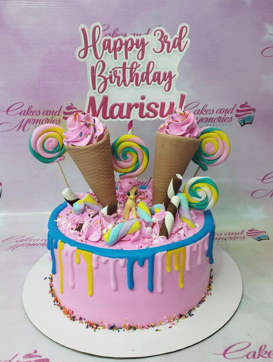 Pink birthday cake with colorful icing drips, ice cream cone toppers, lollipops, and a My Little Pony figurine.