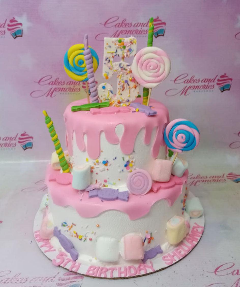 Two-tier white cake with pink icing drips, colorful lollipops, marshmallows, and a large sprinkle-covered number five topper.