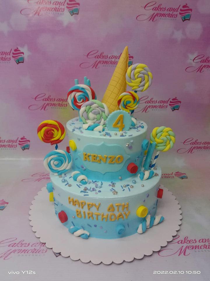 Two-tier light blue candy-themed birthday cake with colorful fondant lollipops, ice cream cone topper, and marshmallow twists.