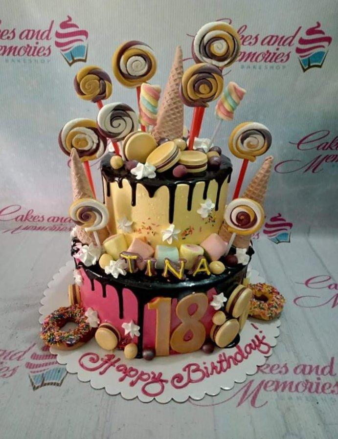 Two-tier candy-themed birthday cake with pink and yellow icing, chocolate drips, lollipops, macarons, and donuts.