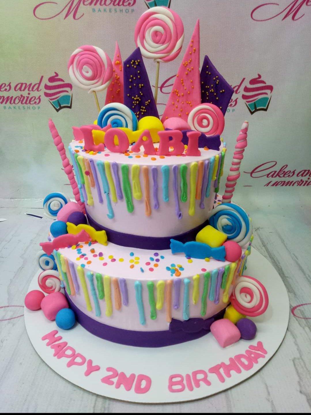 Colorful 2-tier candy themed birthday cake with rainbow drips, fondant lollipops, and pink gumpaste letters for Loabi's 2nd birthday.