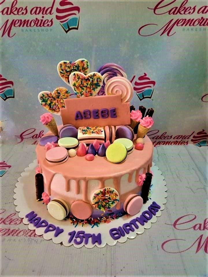 Pink and purple candy-themed birthday cake with macarons, ice cream cones, and heart-shaped toppers with sprinkles.