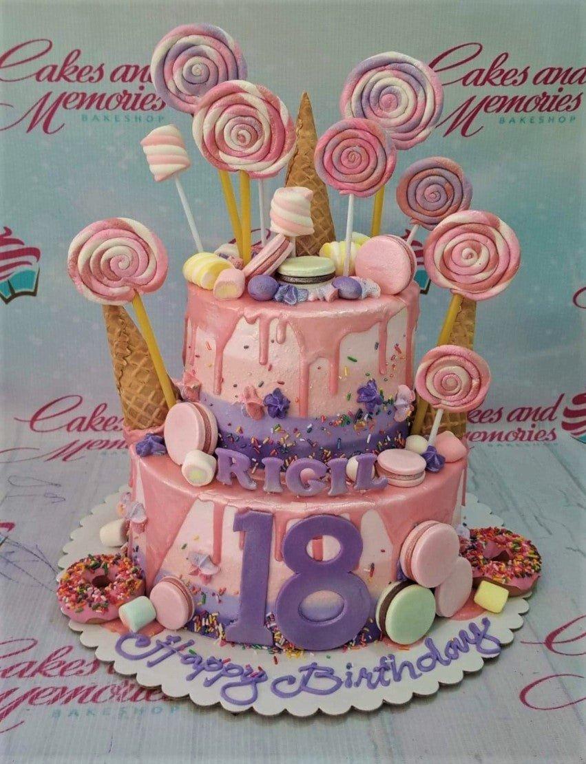 Two-tier pink and purple candy-themed birthday cake with lollipops, macarons, donuts, and a large purple number 18 topper.