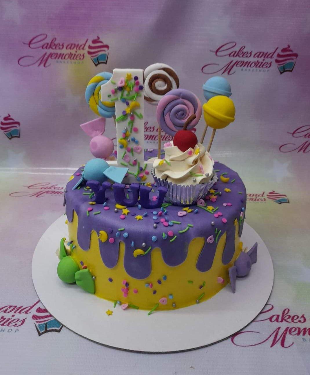 Yellow fondant cake with purple drip, topped with a large number one, a cupcake, and colorful candy-themed fondant toppers.