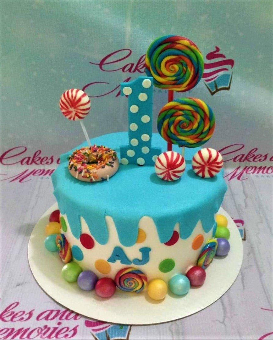 Colorful candy-themed first birthday cake with a blue fondant drip, lollipops, a donut, and a large number one topper.