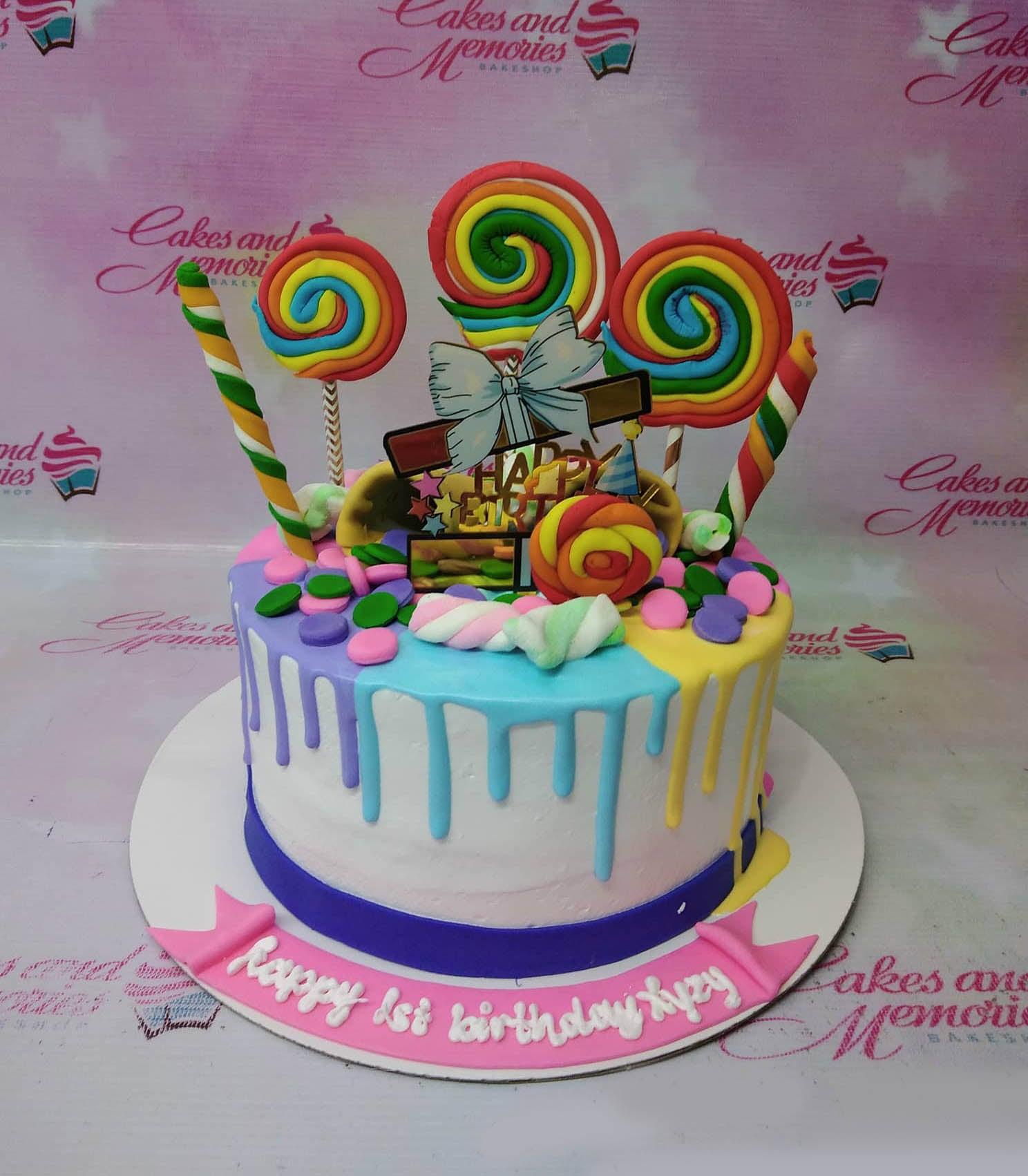 Colorful candy-themed birthday cake with rainbow lollipops, marshmallows, and multi-colored icing drips on a white base.