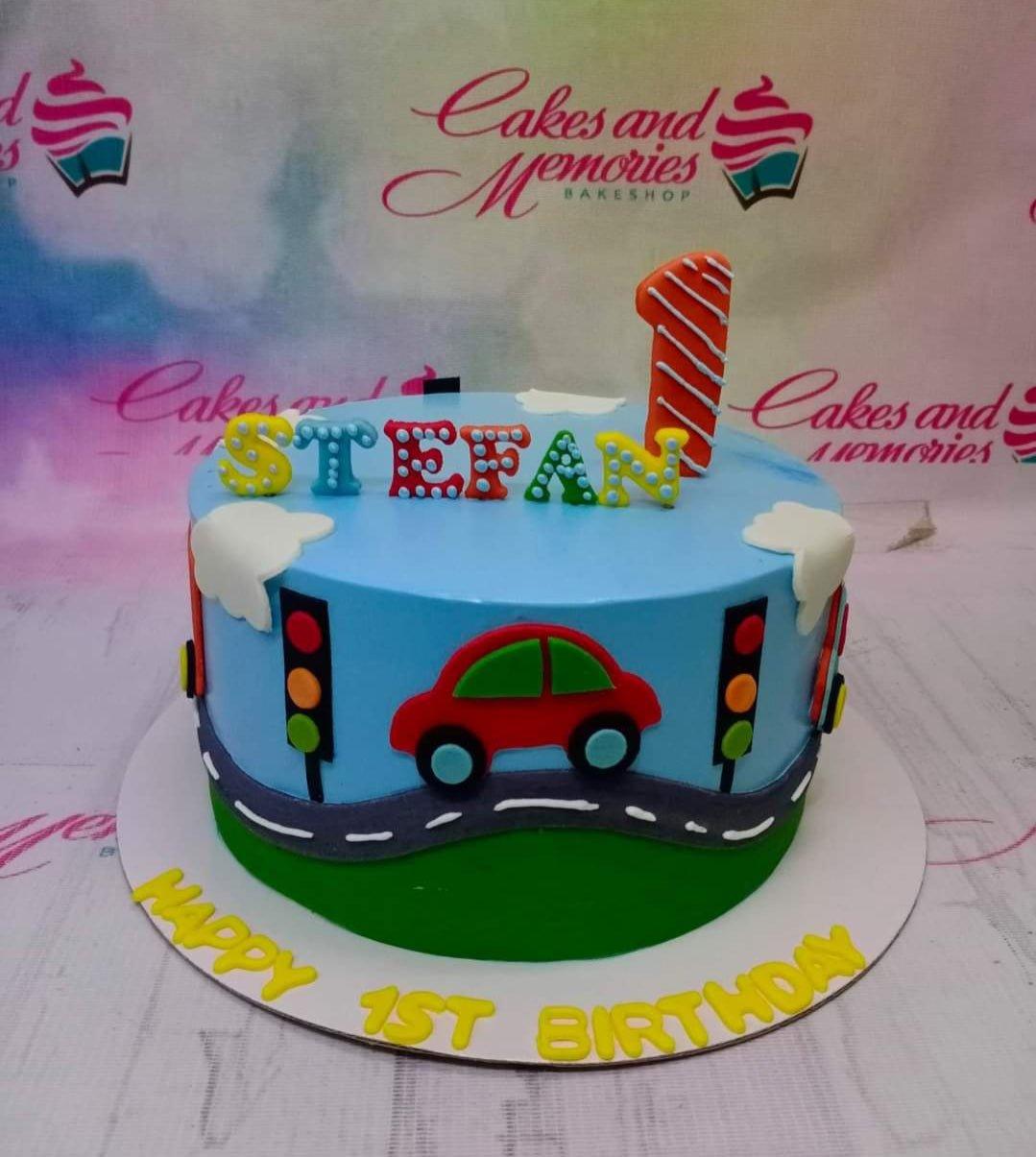 Light blue car themed birthday cake with a red car, traffic lights, and colorful gumpaste letters for Stefan's 1st birthday.