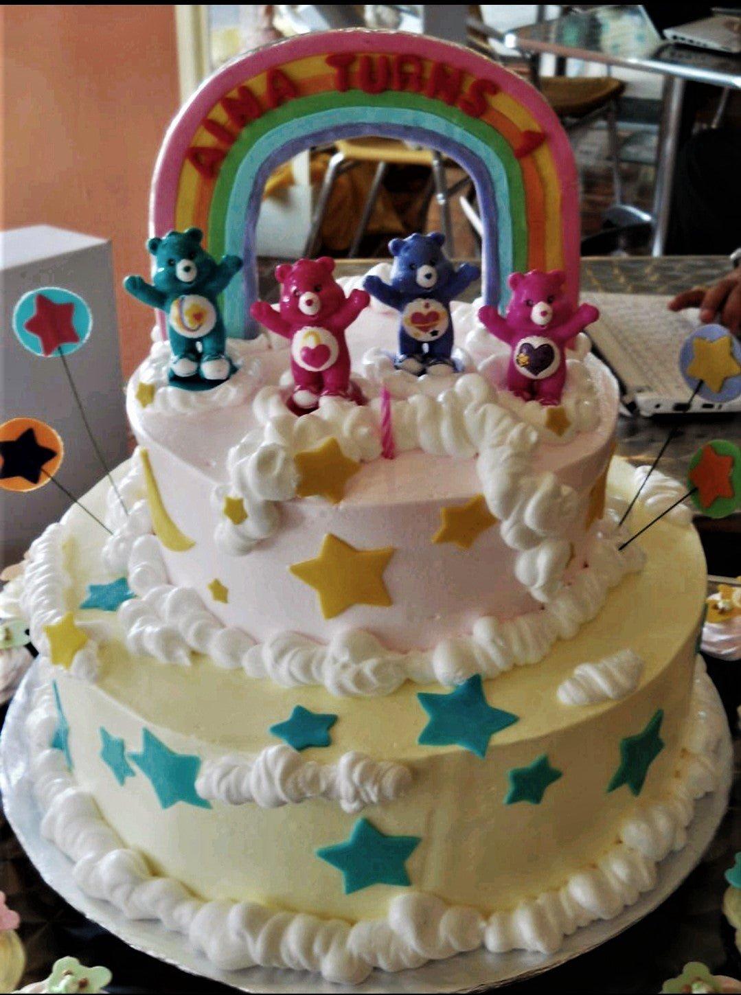 Two-tier Care Bears birthday cake with a rainbow topper, fondant stars, and white icing clouds on pink and yellow tiers.