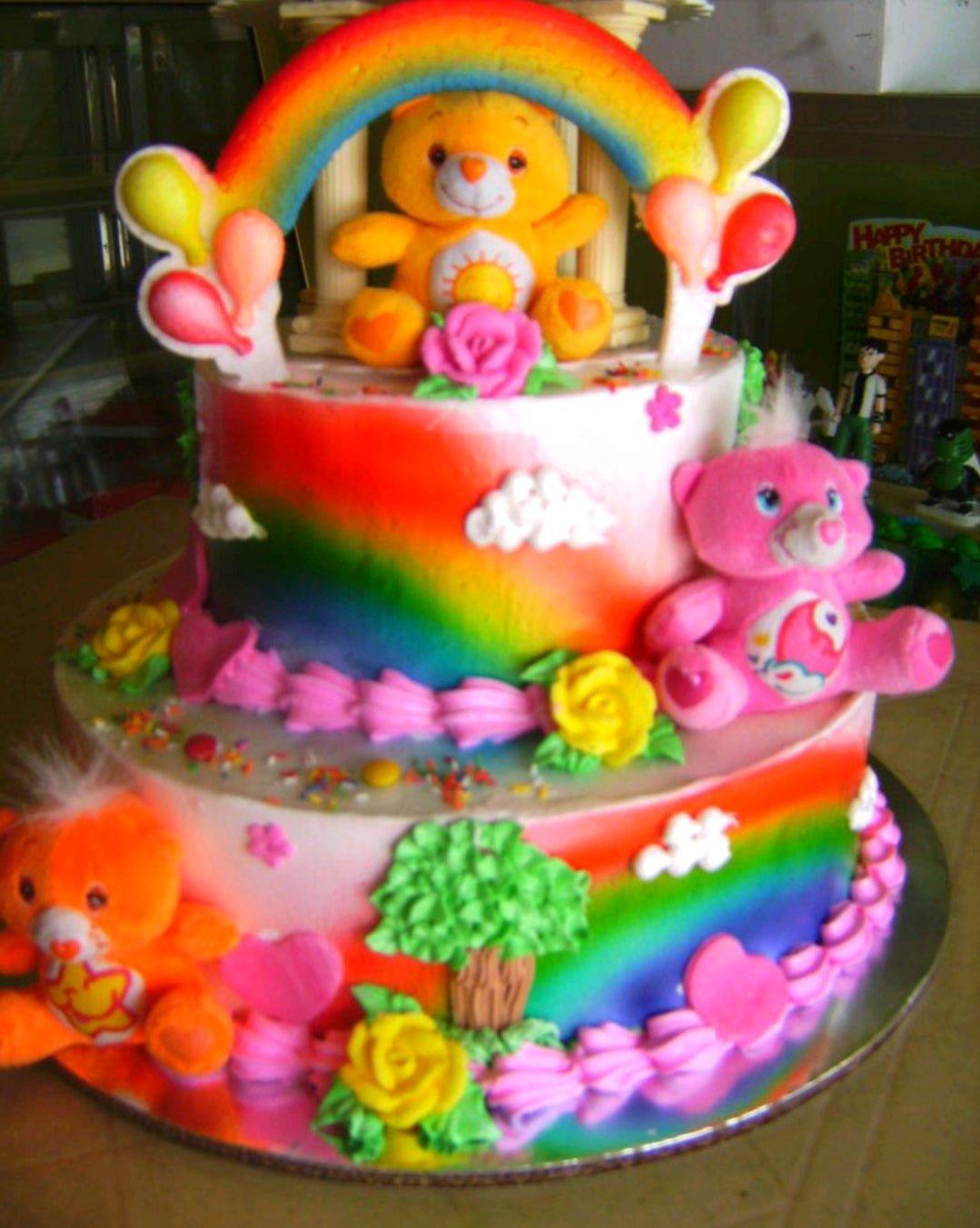 Two-tier rainbow Care Bears cake with orange and pink plush-style toppers, fondant rainbow, and colorful icing rosettes.