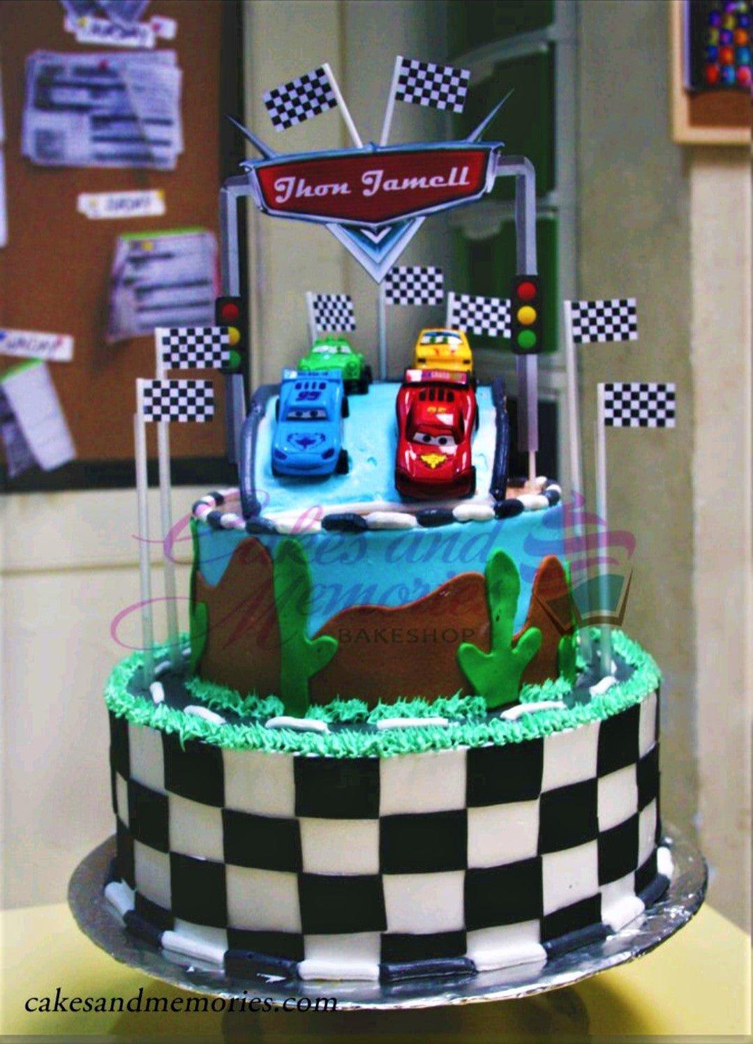 Two-tier Cars themed racing cake with checkered pattern, toy cars, and checkered flags on a blue and green landscape background.