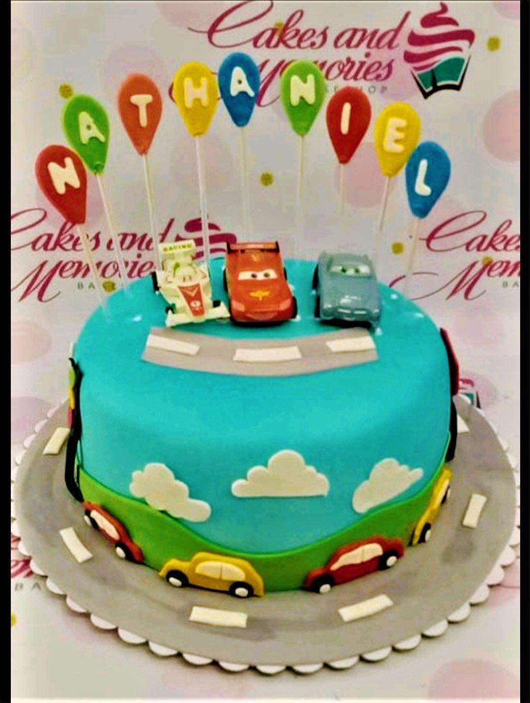 Blue fondant Cars themed birthday cake with toy cars on a road and balloon name toppers for Nathaniel.