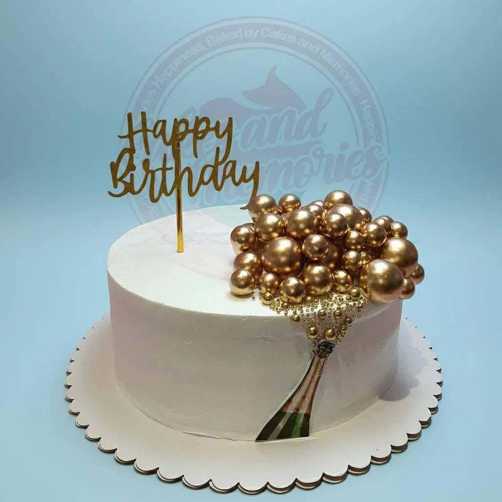A white 1 Tier cake featuring a gold 'Happy Birthday' topper, gold decorative spheres, a champagne bottle printout, and gold sugar beads.