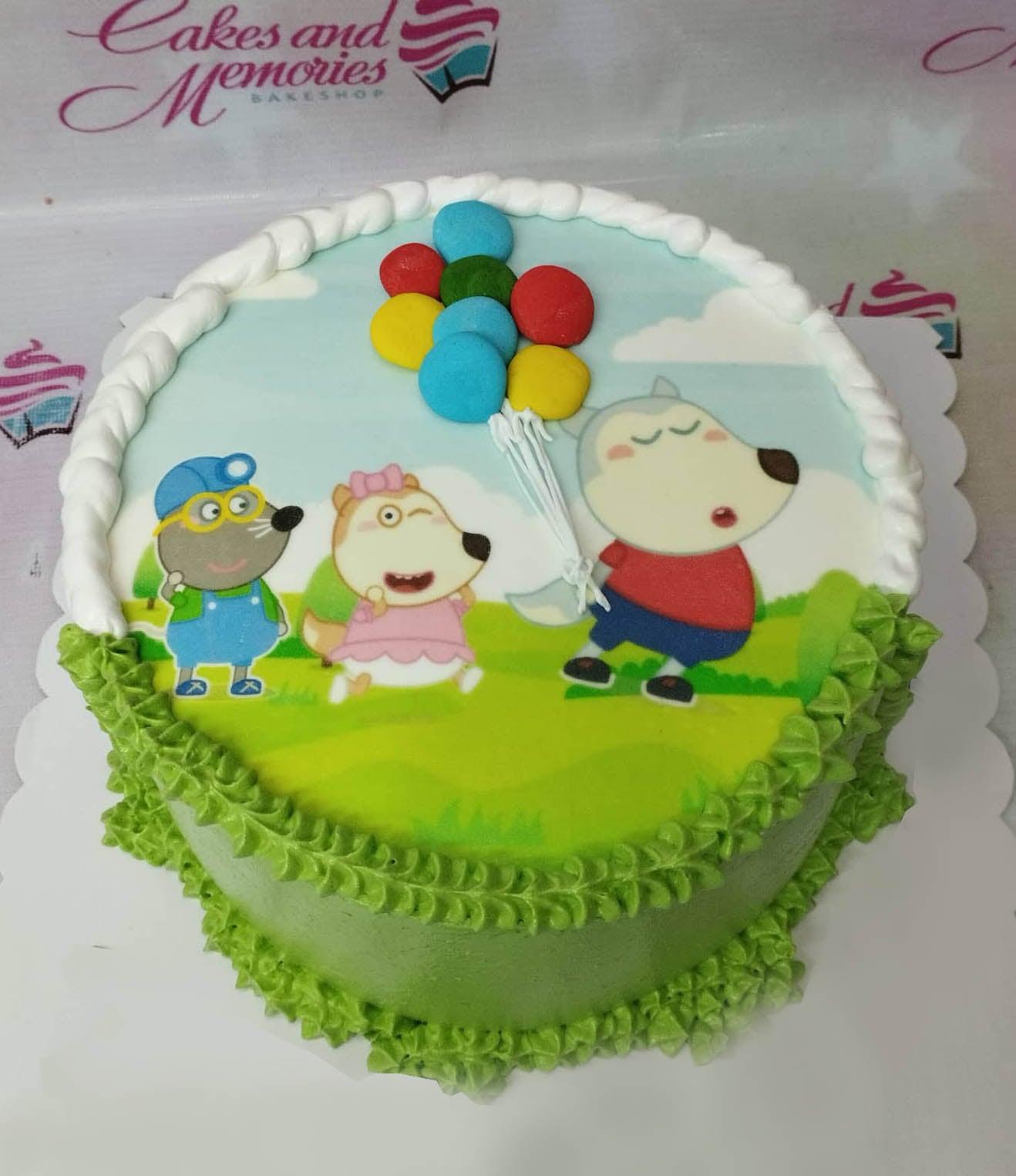 Round green cake with a printed character scene on top featuring a wolf, a dog, and a mouse with colorful balloons.