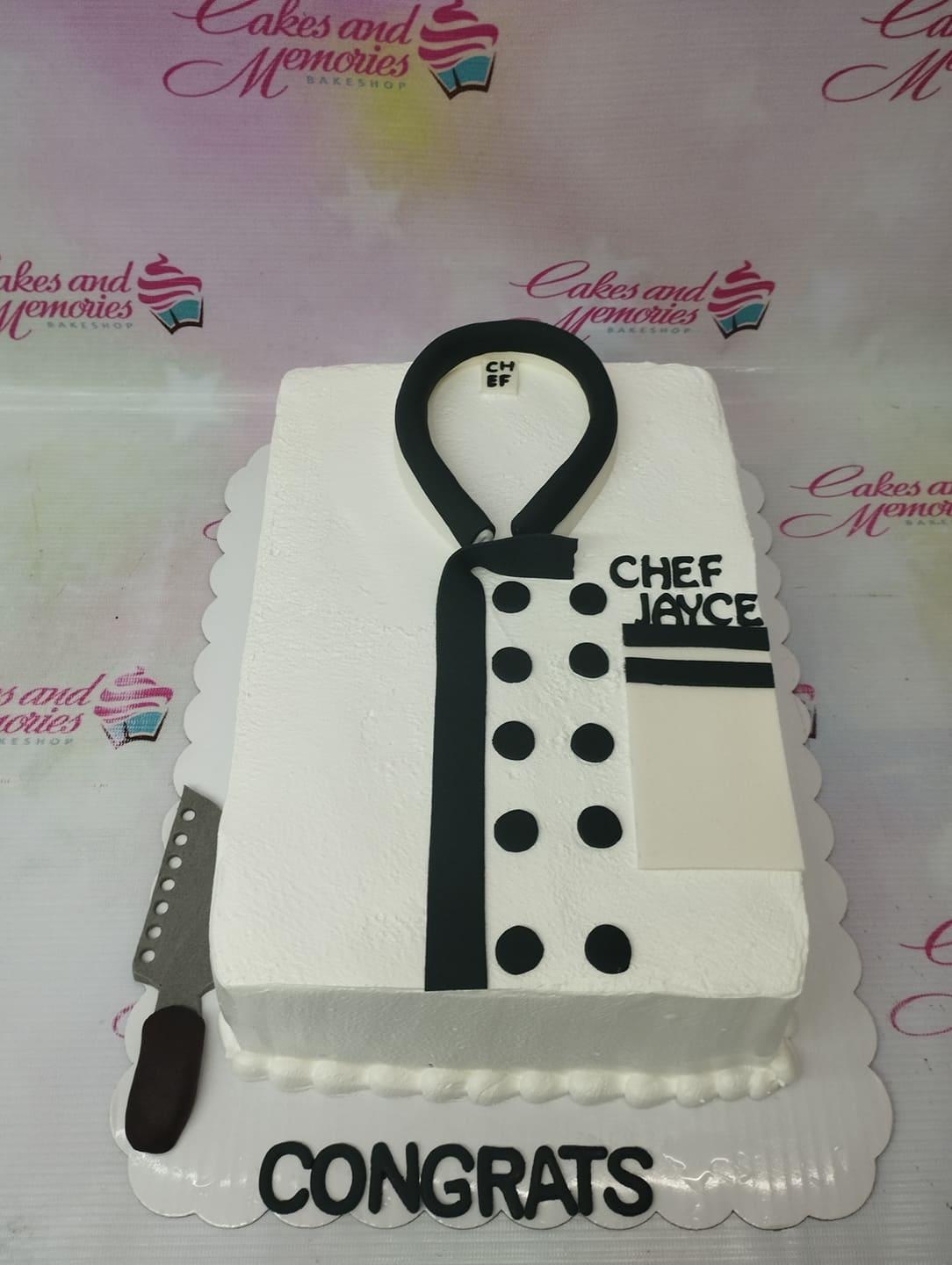 White rectangle chef uniform cake with black fondant buttons, tie, and a gumpaste knife on the side.