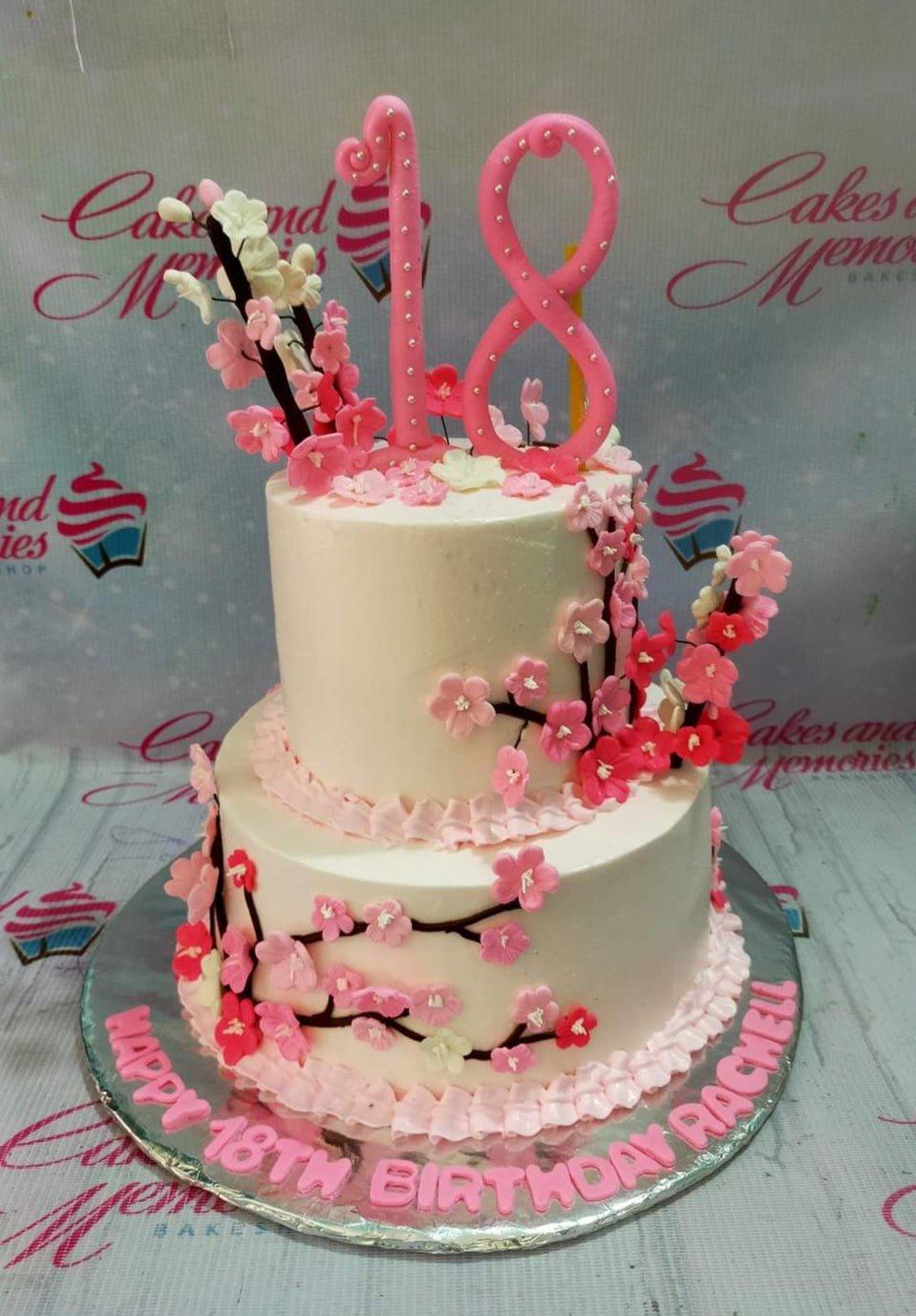 Two-tier white soft icing cake decorated with pink cherry blossom branches and a large pink number 18 topper.
