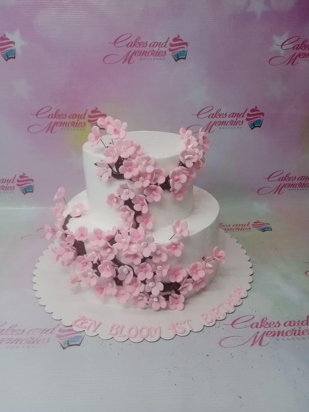 Elegant two-tier white cake decorated with cascading pink cherry blossom flowers and brown branches, featuring a birthday message on the base.