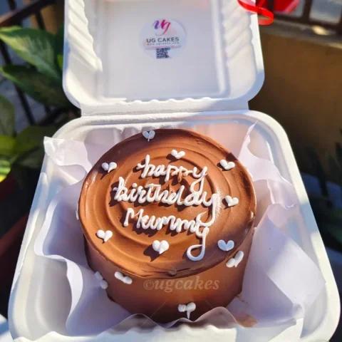 Chocolate bento cake with brown icing, white piped hearts, and 'Happy Birthday Mummy' message in a white takeout container.