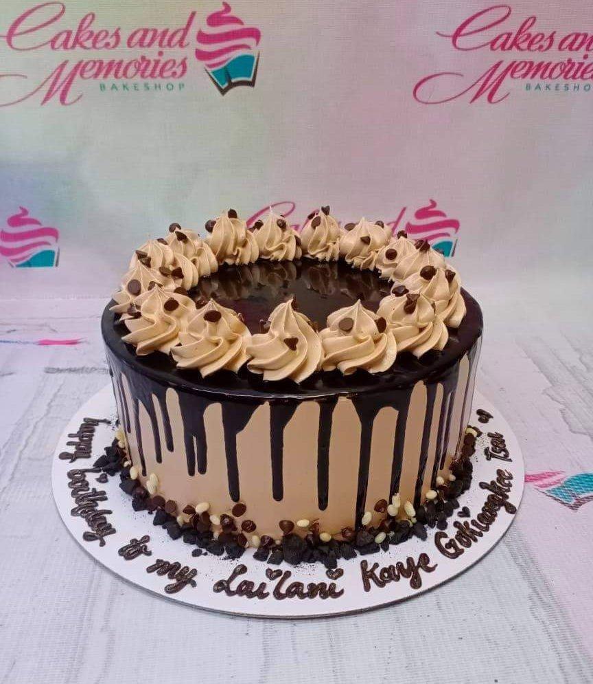 Chocolate drip cake with tan icing rosettes and chocolate chips on a white base board with a birthday message.