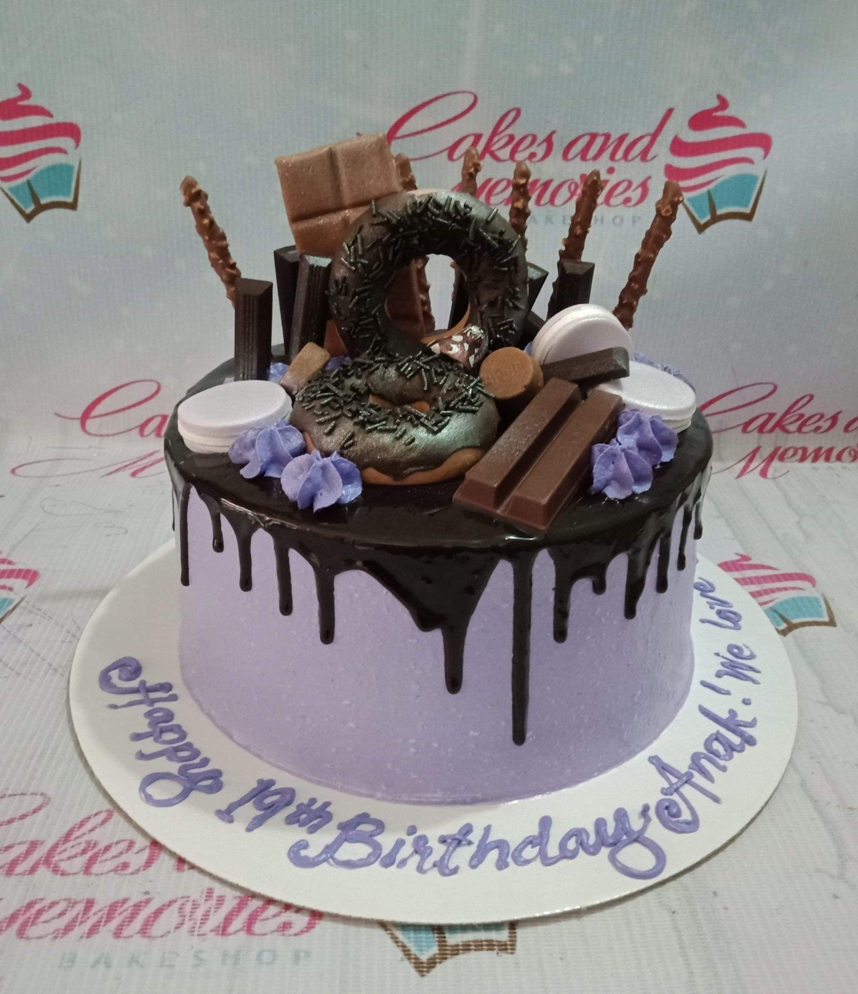 Purple birthday cake with chocolate drip, topped with donuts, chocolate bars, and purple icing rosettes.