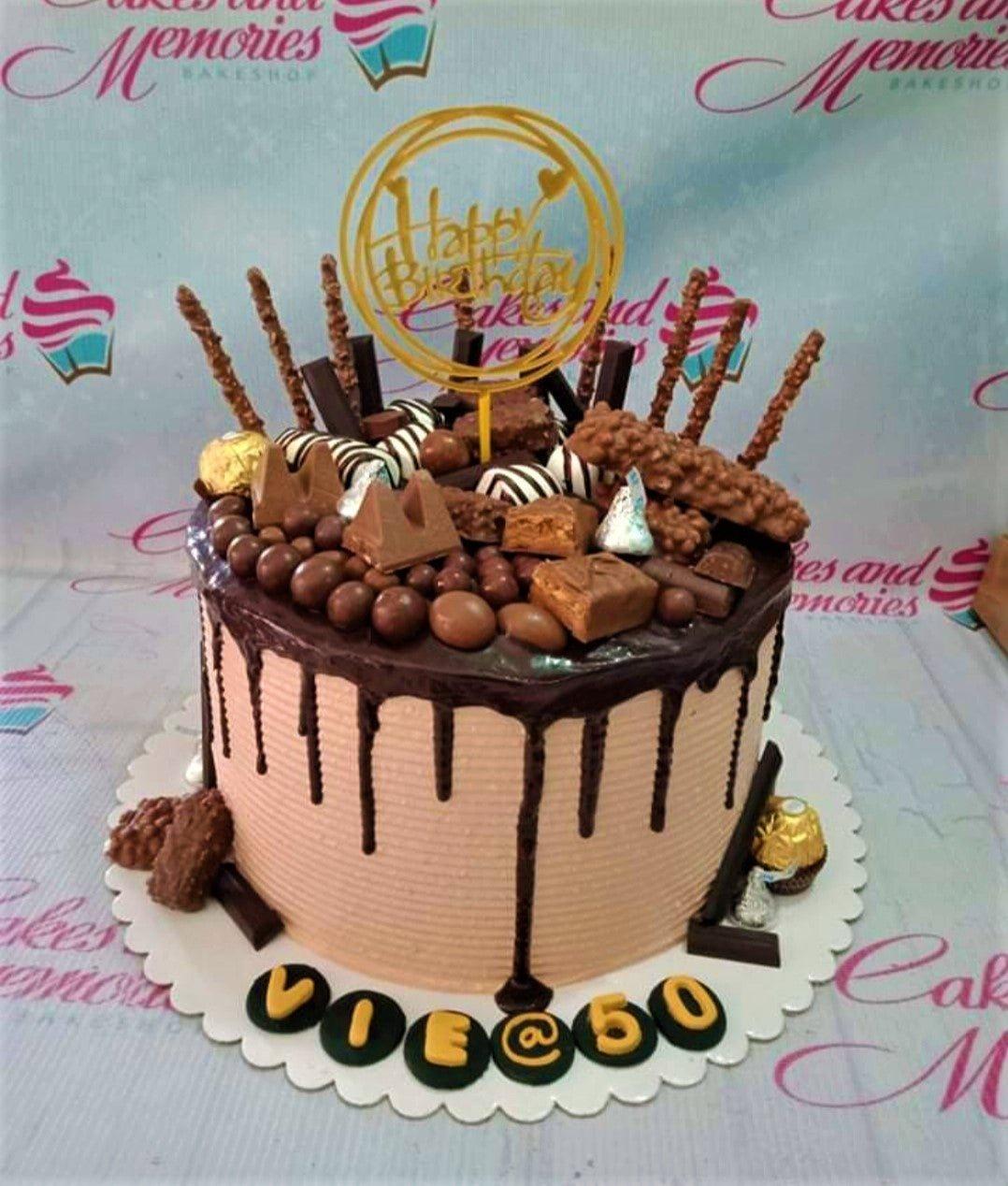 Chocolate drip cake with assorted chocolate bars, Ferrero Rocher, and a gold Happy Birthday topper on light pink icing.