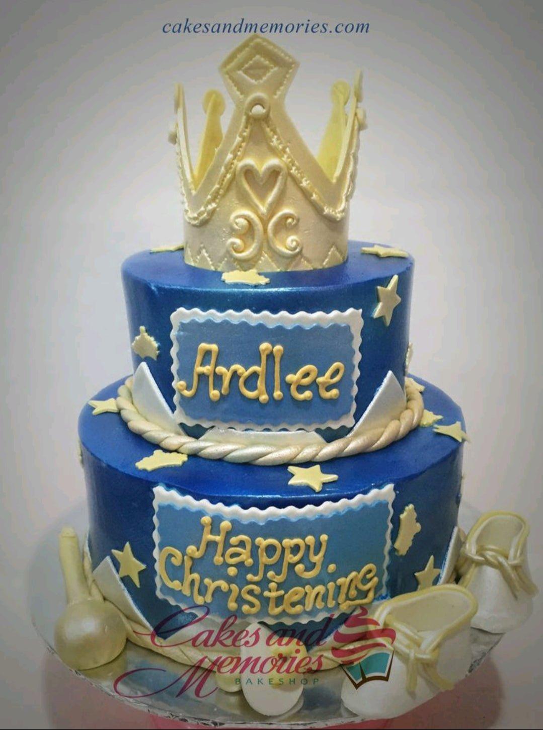 Two-tier royal blue fondant cake with a large gold crown topper, white baby shoes, and gold stars for a christening.