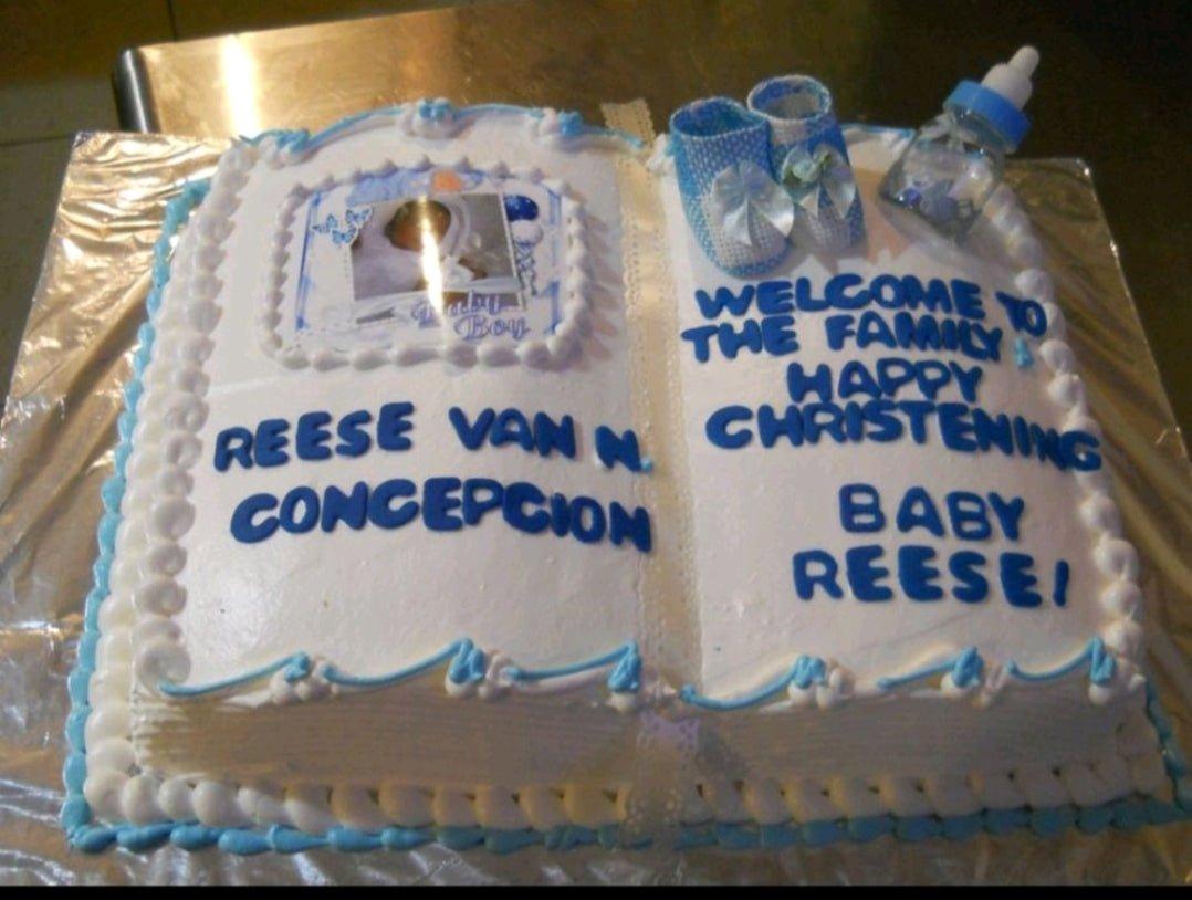 White rectangle book-shaped christening cake with blue icing borders, a baby photo printout, blue baby booties, and a feeding bottle topper.